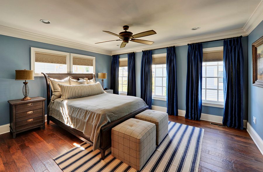  A bedroom with a bed, two windows with dark blue curtains, a brown dresser, a striped rug, a ceiling fan, and an eye mask and white noise machine on the nightstand.